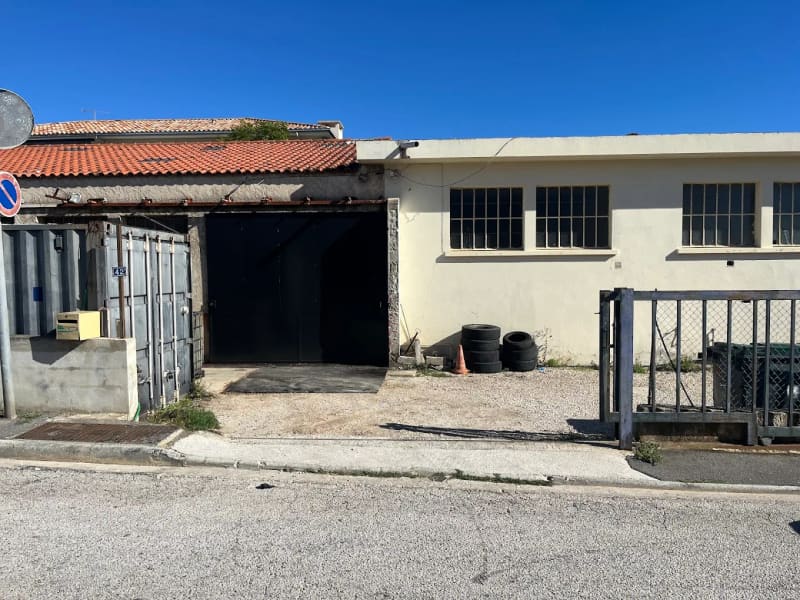 Façade du garage Tout pour l'Auto 83 vue de la rue : grande porte sombre sous un toit de tuiles, bâtiment clair aux fenêtres à petits carreaux, pneus empilés et portail métallique